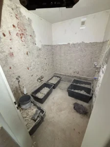 Raum mit Baustelle, abblätterndem Putz, schwarzen Behältern voller Schutt, Eimer am Boden.