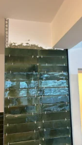 Dunkelgrüne Fliesenwand mit Abstandshaltern, unverputztem Mörtel oben, Metallschiene links.