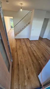 Eingangsbereich mit Holzboden, Treppe mit weißem Geländer und großem Fenster im Hintergrund.