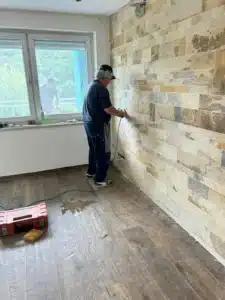 Handwerker bearbeitet Innenwand mit Elektrowerkzeug, Person draußen auf Balkon sichtbar.