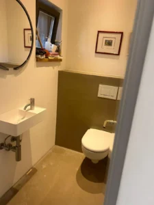 Kleines Badezimmer mit Waschbecken, Rundspiegel, WC, Fenster und hell gestrichenen Wänden.