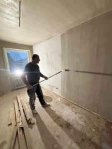 Mann streicht Wand mit Farbrolle in einem unfertigen Raum mit Holzstücken und Fenster.