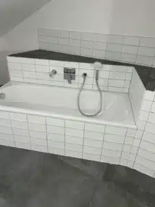Moderne Badewanne mit weißen Fliesen, Wasserhahn, Handbrause, dunkler Boden.