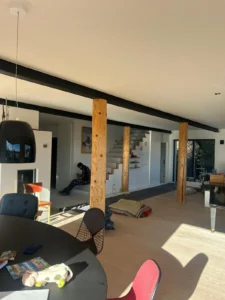 Moderner Wohnraum mit Holzsäulen, rundem Tisch, Decken, Person bei Treppe, Sonnenlicht.