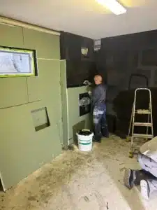 Person renoviert Raum mit teilweise schwarzen Wänden, grüner Trockenbauwand, Leiter und Eimer.