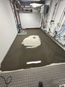 Technikraum mit Rohren, Abfluss und teilweise mit Flüssigkeit bedecktem Boden.